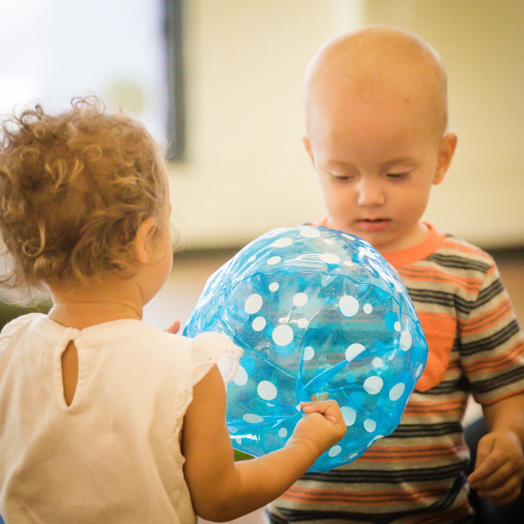 Two toddlers with one offering the other a blue translucent beach ball at RIE® Parent-Infant Guidance™ Class at RIE Pasadena Center