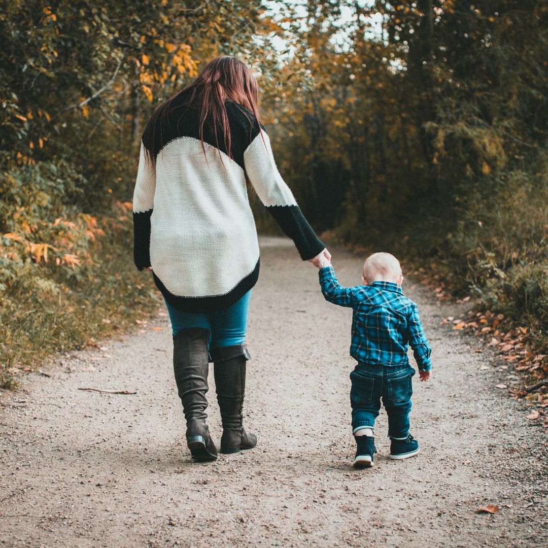 Woman walking and holding hands with a toddler on a gravel path lined with trees and the trees are just beginning to turn colors in the fall.