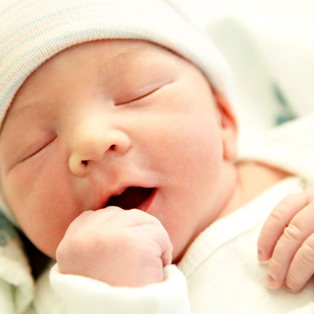 newborn baby in hat with eyes closed with fist coming close to mouth.